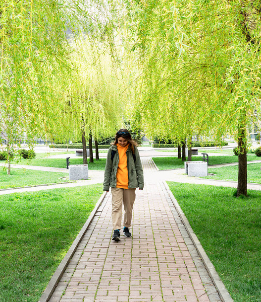 若い緑の春の葉で柳を泣いての路地に沿って歩くバックパックと笑顔の若い女性観光客 - 写真・画像