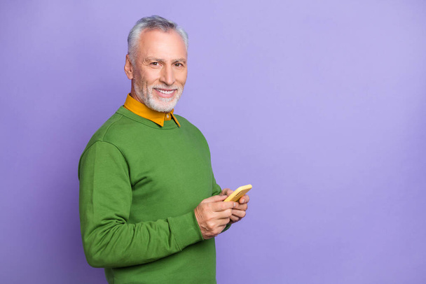 Photo of cheerful retired man wear green sweater writing modern device empty space isolated violet color background - Fotoğraf, Görsel