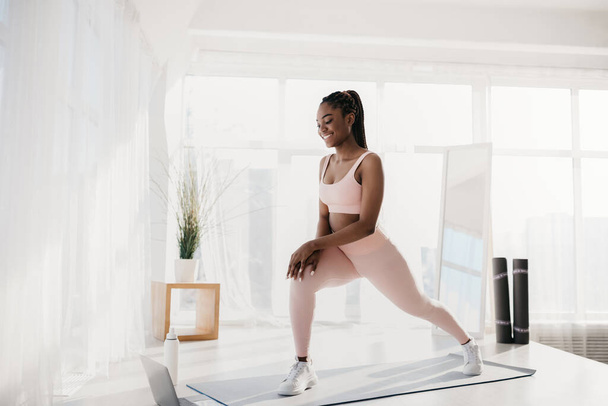 Millennial black woman making lunges near laptop, working out to online sports tutorial at home, free space - Zdjęcie, obraz