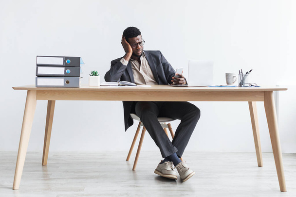 Stres mladý černý podnikatel pomocí smartphone a notebook, sedí za stolem, dělat chyby v obchodním projektu - Fotografie, Obrázek
