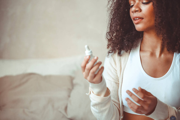 Szépség és bőr koncepció. Vágott fotó afro-amerikai fiatal női modell holding palack hidratáló esszencia szérum ül ellen elmosódott modern lakás belső háttér - Fotó, kép