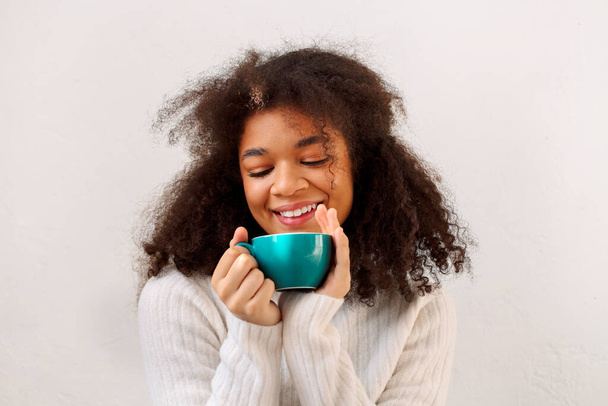 Feliz complacido encantado rizado chica afroamericana con los ojos cerrados disfrutando de la taza de té y relajarse en la fría temporada de invierno, usando suéter blanco de punto cálido, aislado en el fondo del estudio blanco. - Foto, imagen