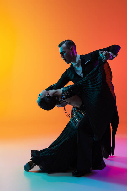 Portrait of young beautiful man and woman dancing ballroom dance isolated over gradient orange pink background in neon light. Beauty, art, sport concept - Valokuva, kuva