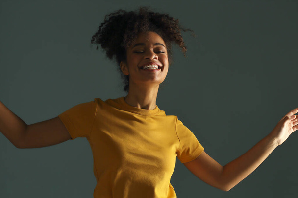 Beautiful happy dark skinned african american ethnicity woman in yellow tshirt raising hand in greeting while looking in camera with pleasant smile, posing against gray green studio wall background - Photo, Image