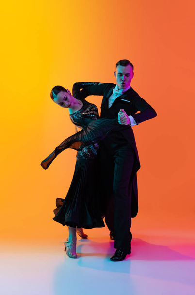 Portrait of young beautiful man and woman dancing ballroom dance isolated over gradient orange pink background in neon light. Beauty, art, sport concept - Valokuva, kuva