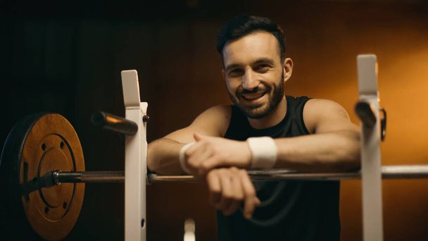 Sportsman smiling at camera near barbell on stand on dark background - Foto, immagini