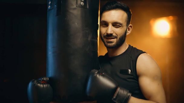Χαμογελώντας αθλητής σε γάντια πυγμαχίας κλείνει το μάτι κοντά τσάντα γροθιά σε σκούρο φόντο - Φωτογραφία, εικόνα