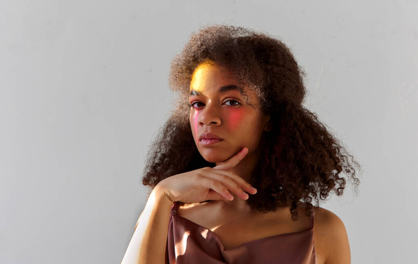 Retrato de estudio creativo del joven modelo afroamericano rizado con maquillaje de arte de color brillante mirando a la cámara, mujer negra con pintura rosa en la mitad de la cara posando aislada sobre fondo gris - Foto, Imagen