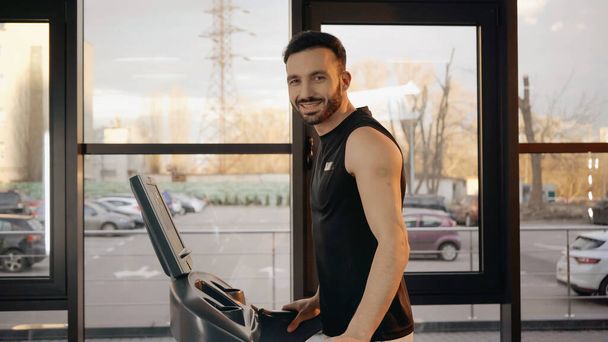Uśmiechnięty sportowiec patrzący w kamerę podczas treningu na bieżni na siłowni  - Zdjęcie, obraz