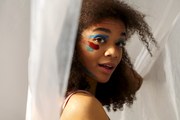 Όμορφη ευτυχισμένη αφροαμερικάνικη γυναίκα εθνικότητας με δημιουργική τέχνη μακιγιάζ κοιτάζοντας πέρα με ευχάριστο χαμόγελο, χαρούμενο μαύρο σγουρό κορίτσι με κηλίδες χρώματος στο πρόσωπο ποζάροντας σε γκρι φόντο στούντιο - Φωτογραφία, εικόνα