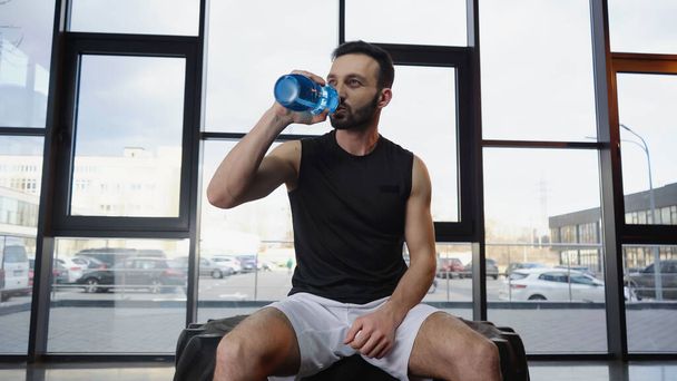 Спортсмен п'є воду, сидячи на шині в спортзалі
  - Фото, зображення