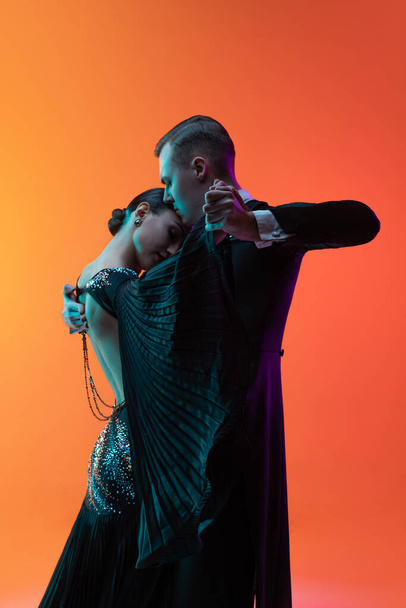 Close-up emotive dancing couple, beautiful man and woman dancing waltz isolated over gradient orange pink background in neon light. Beauty, art, sport concept - Foto, immagini