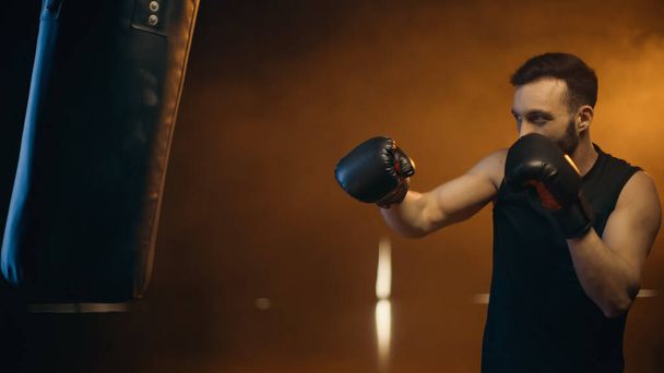 Sportsman in boxing gloves training with punch bag on dark background - Zdjęcie, obraz