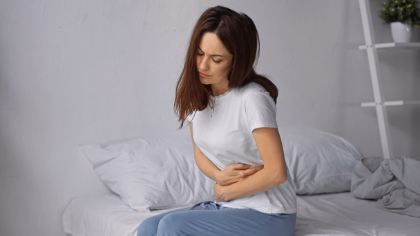 woman sitting on bed and hugging belly while feeling abdominal pain - Photo, Image