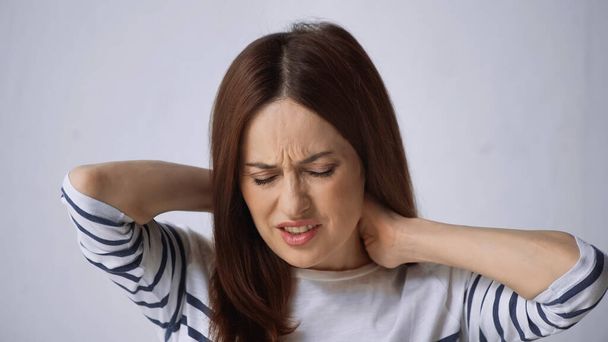μελαχρινή γυναίκα με κλειστά μάτια αγγίζει το λαιμό και συνοφρύωμα από τον πόνο σε γκρι φόντο - Φωτογραφία, εικόνα