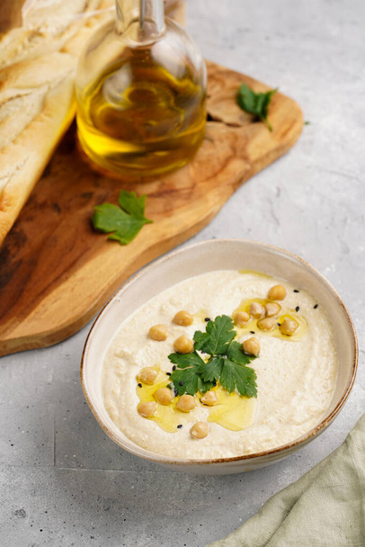 Hummus-Dip mit Kichererbsen und Petersilie in Keramikschale auf Betonoberfläche mit gerösteten Baguettescheiben und Olivenöl auf Holzbrett - Foto, Bild