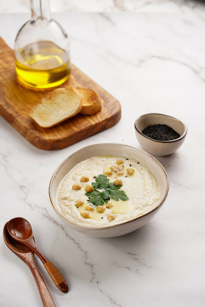 Hummus-Dip mit Kichererbsen und Petersilie in Keramikschale auf Marmoroberfläche mit gerösteten Baguettescheiben und Olivenöl auf Holzbrett - Foto, Bild