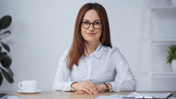 pleased businesswoman in eyeglasses sitting at workplace and looking at camera - Valokuva, kuva