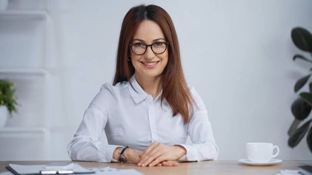 brunette businesswoman in eyeglasses sitting at workplace and smiling at camera - Fotoğraf, Görsel