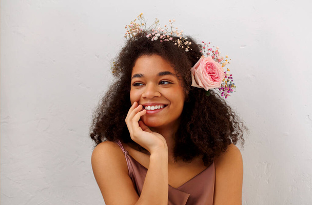 Kıvırcık saçlı, gri arkaplanda izole edilmiş taze pembe gül çiçekli, gülümseyen Afro-Amerikan genç kadının bahar portresini kapatın. Afrikalı kadınların doğal güzelliği - Fotoğraf, Görsel
