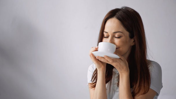 pleased woman with closed eyes enjoying morning coffee on grey background - Photo, Image