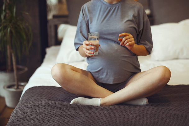 Ergänzungsmittel während der Schwangerschaft. Junge, glückliche Schwangere, die pränatale Vitamine nimmt, ein Glas Wasser und Tabletten in der Hand hält, während sie zu Hause im Bett liegt. Werdende Mutter beginnt jeden Tag mit Folsäure - Foto, Bild