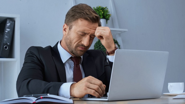 exhausted businessman sitting with closed eyes near laptop and suffering from headache - Foto, Imagen