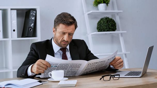 homem de negócios em terno preto lendo notícias econômicas perto de xícara de café e laptop - Foto, Imagem