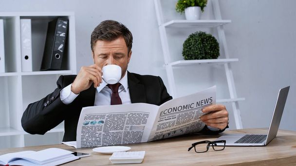 ビジネスマンはオフィスのノートパソコンの近くでコーヒーを飲み新聞を読む - 写真・画像