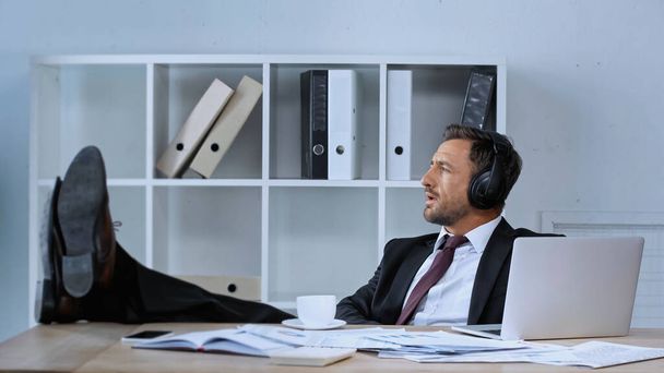 hombre de negocios con auriculares sentados con piernas en el escritorio mientras escucha música durante el descanso del café - Foto, Imagen