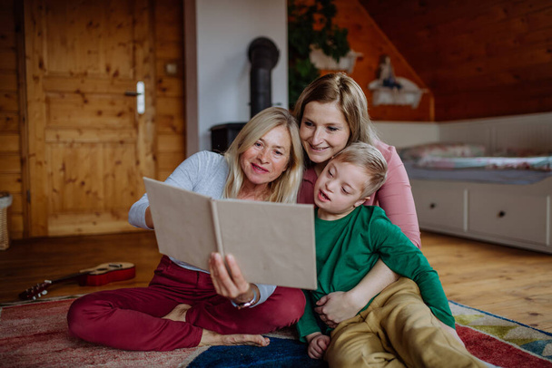 Boy with Down syndrome with his mother and grandmother looking at family photo album at home. - Zdjęcie, obraz