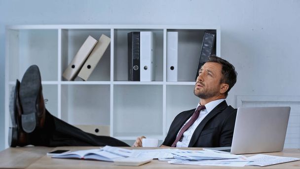 man in suit sitting with legs on desk while resting during coffee break in office - Photo, image