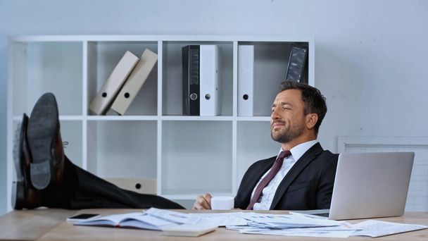 pleased businessman sitting with legs on desk while resting during coffee break - Photo, image
