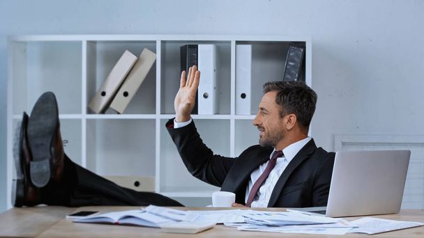 side view of happy man waving hand while sitting with legs on desk at workplace - Photo, Image