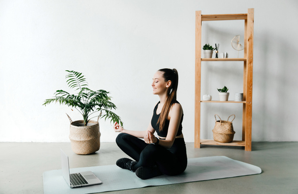 Koncept on-line školení. Mladá štíhlá žena cvičí jógu doma v online lekcích, sedí na podložce, notebook před ní - Fotografie, Obrázek