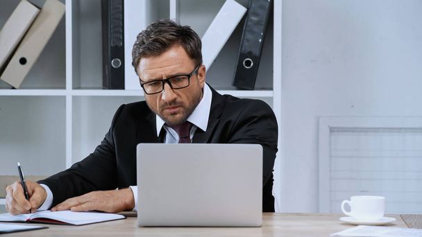 businessman in eyeglasses writing in notebook while looking at laptop in office - Foto, Imagem