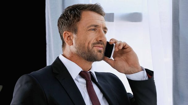 zakenman in pak glimlachen terwijl praten op mobiele telefoon op kantoor - Foto, afbeelding