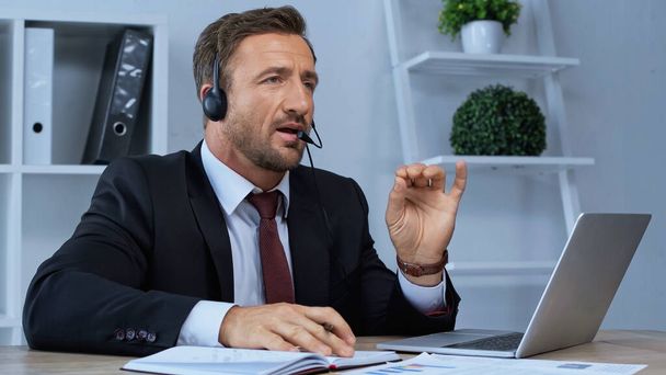 man in headset talking and gesturing near laptop at workplace - Фото, зображення