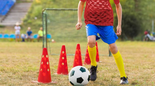 10代の少年は学校のフィールドでサッカーのトレーニングに出席します。物理的な教育のクラスの若者。子供のためのサッカー練習。コーンスラロームパッチでボールを実行している選手 - 写真・画像