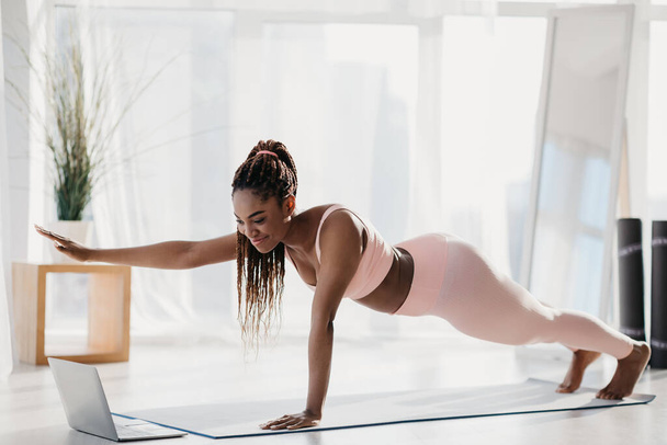 Fit young black woman standing in plank with outstretched arm, following online video tutorial on laptop at home - Foto, Imagen