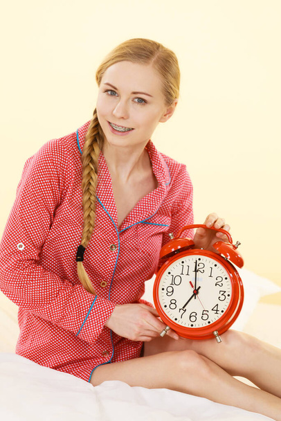 Sleepy young woman wearing cute pink pajamas holding big red old fashioned clock showing sleep time. - Photo, Image