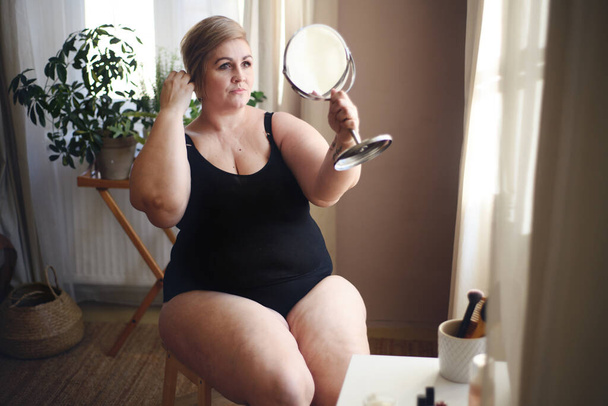 Overweight woman sitting and looking at mirror at home, selfcare concept. - Photo, image