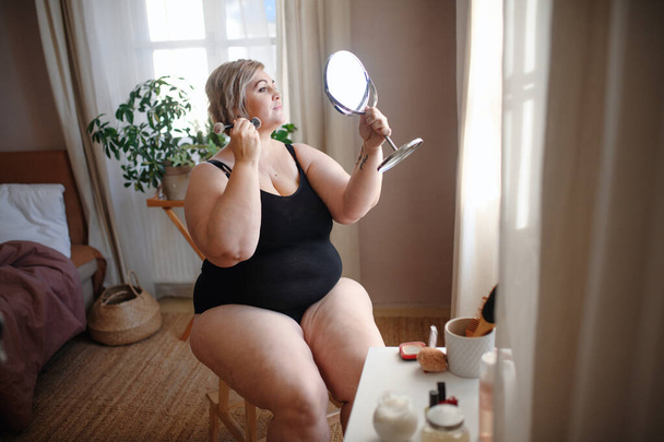 Overweight woman sitting and looking at mirror and applying make up at home, selfcare concept. - Fotó, kép