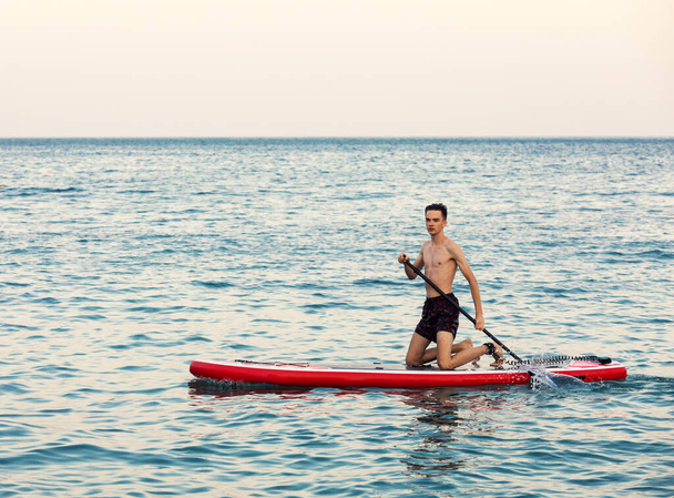 Teenager boy learning to paddle on a SUP board in the sea kneeling - 写真・画像
