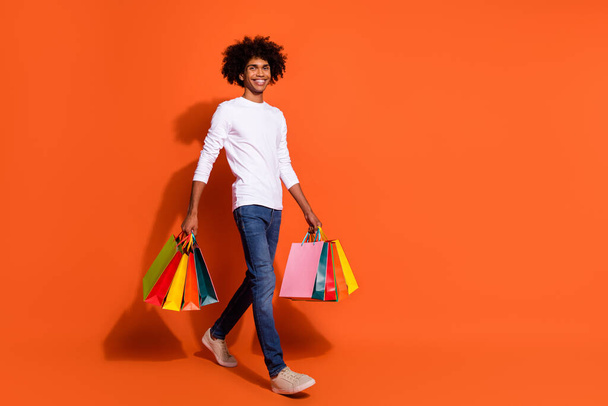 Photo of attractive pretty guy dressed white shirt carrying shoppers walking empty space isolated orange color background. - Φωτογραφία, εικόνα