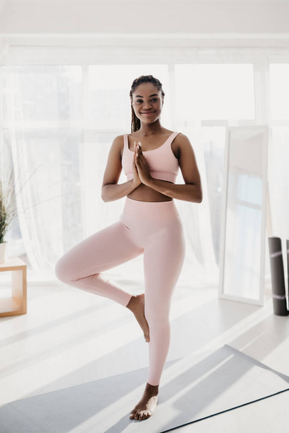 Glückliche junge schwarze Frau in Sportkleidung steht in Baumpose und praktiziert Yoga auf Matte zu Hause im Fitnessstudio, in voller Länge - Foto, Bild