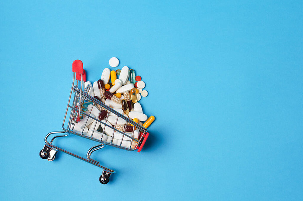 Shopping cart trolley with assorted medicine pills on blue medical background. Concept: idea for online pharmacy, drug delivery, pharmaceutical company business concept. Handful of pills. Copy space - Фото, изображение