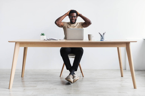 Шокированный черный мужчина сидит за столом с ноутбуком, хватается за голову в ужасе, делает ошибку в проекте, пропускает дедлайн - Фото, изображение