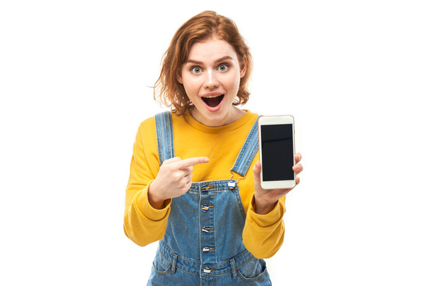 Shocked joyful redhead girl points finger at smartphone blank screen in yellow clothes isolated on white studio background - Valokuva, kuva