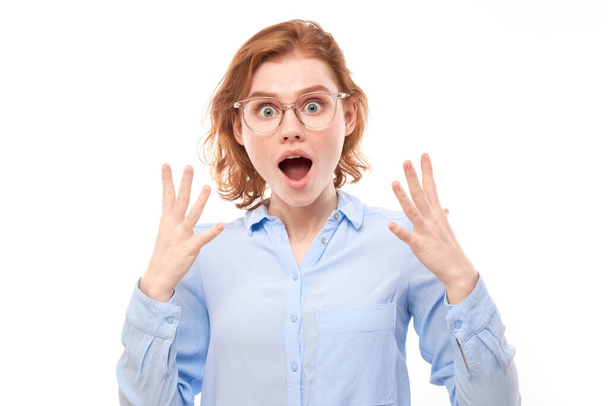 Shocked face red-haired girl in business shirt looks surprised with open mouth spreads hands on white studio background - Foto, afbeelding